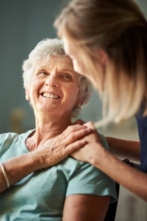 A caregiver rests a reassuring hand on a smiling woman's shoulder at home