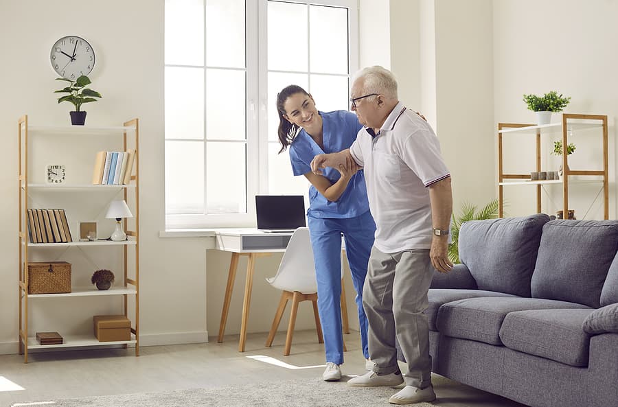 A caregiver walks arm in arm with a man through his living room